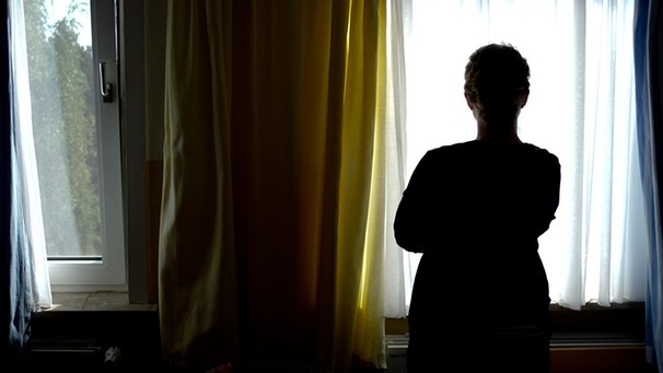 Frau steht mit dem Rücken zum Betrachter in einem dunklen Zimmer und blickt durch eine Gardine aus dem Fenster | Bild: dpa-Bildfunk/Peter Steffen