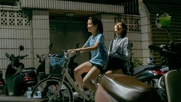 Zwei junge Frauen fahren auf einem Fahrrad durch eine kleine Straße, irgendwo in Taiwan. Eine Frau fährt das Fahrrad, die andere sitzt auf dem Gepäckträger und wirkt verträumt. | Bild: Fragrance Of The First Flower/PORTICO MEDIA ORIGINALS PRODUCTION