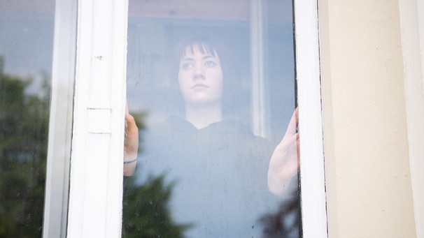 Eine Jugendliche steht in ihrem Zimmer am Fenster, in dessen Scheiben sich Wolken spiegeln (gestellte Szene). | Bild: Paul Zinken/dpa