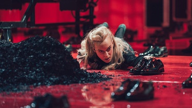 Eine blonde Frau liegt bäuchlings auf rotem Bühnenboden zwischen schwarzen Lackschuhpaaren und Erdhaufen  | Bild: Denis Kuhnert/ Münchner Kammerspiele