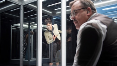 Theaterszene: Ein Mann mit Brille steht in einem Glaskasten, eine kurzhaarige Frau beobachtet ihn | Bild: Heinz Holzmann