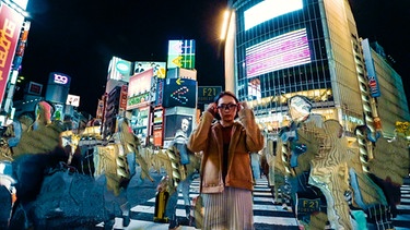 Eine junge Frau steht inmitten einer erleuchteten hochmodernen Großstadt, sie trägt eine Brille, die nach eienm technischen Gerät aussieht, während die Menschen um sie herum als stark verzerrte Kunstfiguren erscheinen | Bild: ars electronica / Terminal Slam