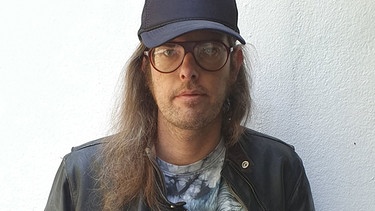 DJ BELP aka Sebastian Schnitzenbaumer blickt, mit Kappe und langen Haaren vor einer hellen Wand stehend, in die Kamera | Bild: Alternative Fakten