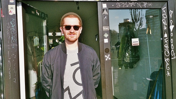 DJ Tom Bauer steht vor einer Tür und blickt mit Sonnenbrille lächelnd in die Sonne | Bild: Alternative Fakten