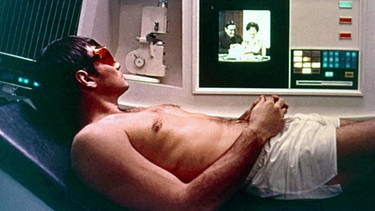Aus dem Film A Space Odyssey - in einem futuristischen Setting liegt ein halbnackter Mann, der TV schaut | Bild: picture alliance/Mary Evans Picture Library