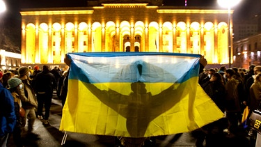 Georgien, Tiflis am 01.03.2022: Ein Demonstrant hält eine ukrainische Fahne vor dem georgischen Parlament während einer Demonstration gegen Russlands Angriff auf die Ukraine hoch. | Bild: Shakh Aivazov/AP/dpa