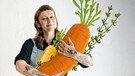 Eine Frau Ende 30 mit tätowierten Armen und Kochschürze umarmt eine Karotte aus Stoff | Bild: Annabell Sievert-Erlinghagen