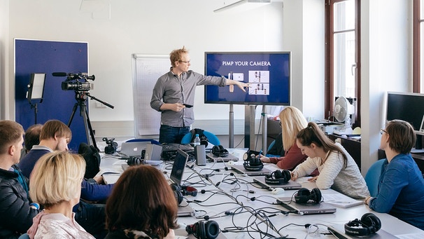 Ein Seminarleiter erklärt einer Gruppe Teilnehmer, wie man eine Kamera benutzt. | Bild: BR/Kimmerzwinger