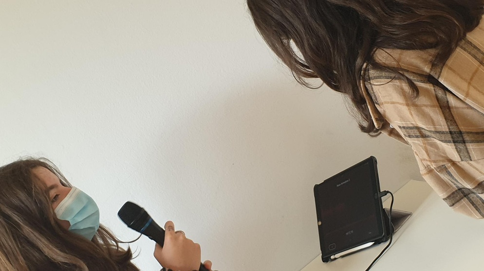 MünchenHören 2021: Ein Mädchen mit Maske hält ein Mikrofon in der Hand, das an ein iPad angeschlossen ist. Ein zweites Mädchen sitzt daneben.  | Bild: BR |Bettina Eisele