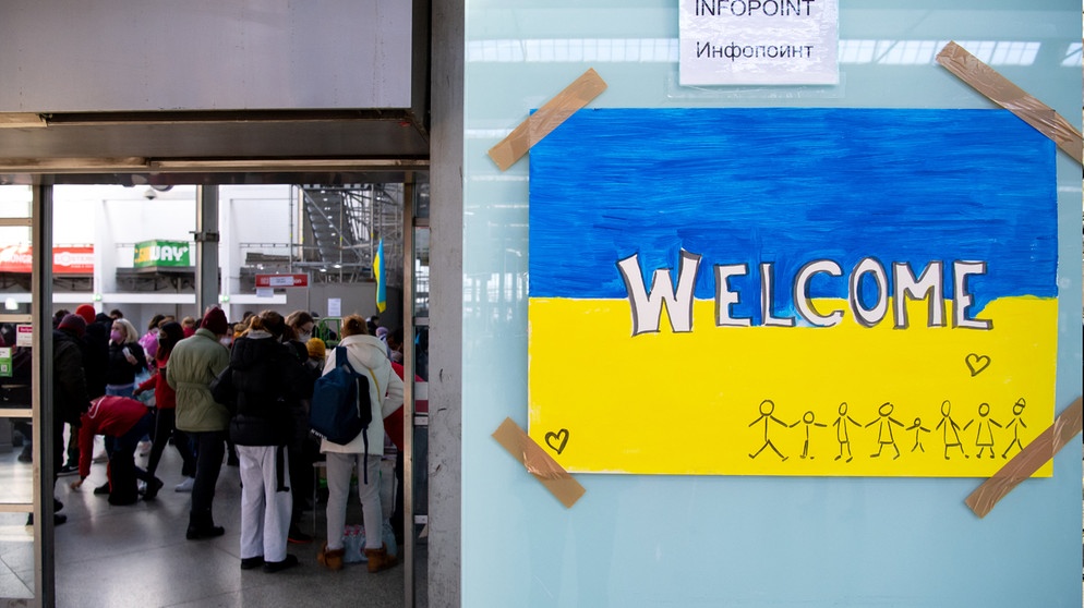 Ein Schild in den Farben der Ukraine mit der Aufschrift "Welcome" ist am Hauptbahnhof zu sehen. Im Hintergrund werden Flüchtlinge aus der Ukraine nach ihrer Ankunft von Mitarbeitern der Caritas und freiwilligen Helfern empfangen.  | Bild: dpa-Bildfunk/Sven Hoppe