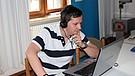 Ein Mann mit Kopfhörern sitzt an einem Tisch und blickt auf einen Laptop. | Bild: BR