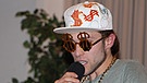 Sebastian Fuchs singt ins Mikro; er trägt ein weißes Käppi mit orangefarbenen Dollarzeichen darauf, eine dicke Goldkette und einen Ring, der drei Finger bedeckt. | Bild: BR