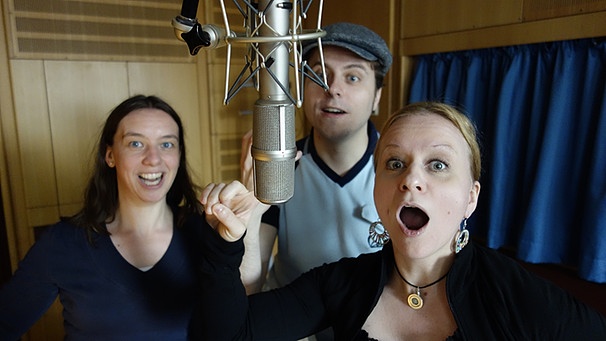 Zwei Frauen und ein Mann stehen im Studio vor einem Mikro mit offenem Mund. | Bild: BR