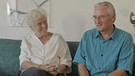 Elke und Hans Martin Schroeder sitzend lächelnd auf dem Sofa in ihrer Wohnung. | Bild: BR