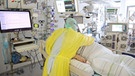 Eine Person liegt im Bett auf einer Intensivstation. Ein Pfleger in Schutzkleidung beugt sich über ihn. Um die beiden stehen viele Geräte unter anderem mit Bildschirmen. | Bild: dpa/Bodo Schakow
