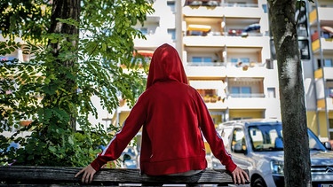 Ein Jugendlicher in roter Kapuzenjacke sitzt auf einem Zaun vor einem Hochhaus. | Bild: BR