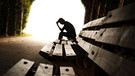 Ein Mensch sitzt zusammengesunken auf einer Bank. Im Vordergrund ist es düster, im Hintergrund scheint ein grelles Licht (Symbolbild) | Bild: stock.adobe.com/hikrcn