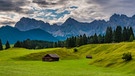 Grüne Buckelwiesen mit drei kleinen Hütten  vorn im Hintergrund die grauen Felsgipfel des Karwendels bei Mittenwald darüber leicht bewölkter Himmel  | Bild: stock.adobe.com/Gina Bromá