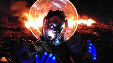 Eine weibliche Person Of Colour mit Dreadlocks, den Kopf in einem Raumfahrerhelm, streckt blau behandschuhte Hände nach oben - Hinter ihr lodert in der Dunkelheit eine horizontale Feuersbrunst | Bild: Münchner Kammerspiele