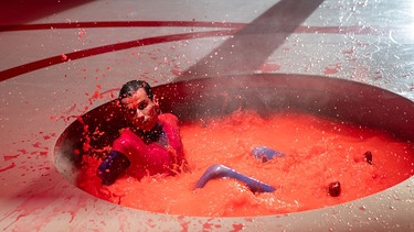 Mann im roten Pullunder sitzt in einem Whirl Pool mit spritzender, roter Flüssigkeit.  | Bild: Münchner Kammerspiele / Krafft Angerer