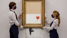 Zwei Angestellte von Sotheby's halten das Banksy-Werk "Love is in the Bin" während einer Versteigerung | Bild: Sotheby's/PA Media/dpa