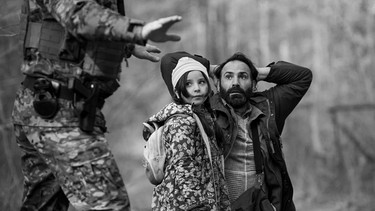 Man sieht einen Mann mit erhobenen Händen sowie  seiner kleinen Tochter vor einem Grenzbeamten niederknien. | Bild: Agata Kubis / Piffl Medien