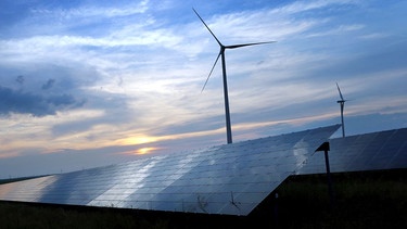 Der mit Wolken überzogene Abendhimmel spiegelt sich auf den Solarzellen einer vor Windrädern stehenden Solarkraftanlage.  | Bild: dpa/ Picture Alliance