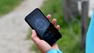 Hand mit Smartphone auf der die Kompass-App zu sehen ist, im Hintergrund ein Wanderweg.  | Bild: picture alliance / BARBARA GINDL / APA / picturedesk.com | BARBARA GINDL