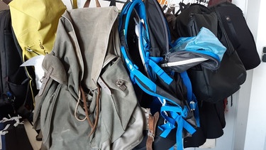 Ein alter grauer und ein neuer blauer Rucksack mit Reparaturbedarf hängen an Kleiderständer, dahinter schwarze Rucksäcke und Taschen und ein gelber Schutzsack    | Bild: Angela Braun/BR