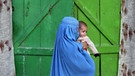 Eine Frau unter einer blauen Burka trägt ein kleines Mädchen und geht an einer Tür vorbei | Bild: picture-alliance / AFP Creative | Ed Jones