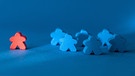Fünf blaue Spielfiguren stehen in einem Kreis eine rote Spielfigur steht etwas abseits in der Nähe einer nicht sichtbaren Lichtquelle. | Bild: picture alliance / Zoonar | Barbara Neveu