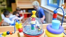 Bunte Spielsachen stehen in einer Kita; im Hintergrund sieht man unscharf eine Erzieherin mit mehreren Kindern. | Bild: picture alliance / Uwe Anspach/dpa | Uwe Anspach