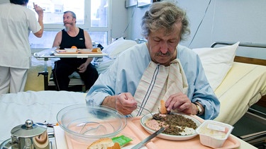Mann sitzt am Rande eines Krankenhausbettes, vor ihm ein Teller Essen.  | Bild: picture-alliance / BSIP/BELMONTE | BELMONTE