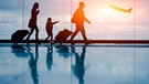 Silhouette einer jungen Familie mit Koffern, im Hintergrund ein startendes Flugzeug | Bild: stock.adobe.com/NicoElNino