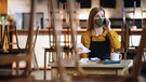 Eine Frau steht mit Maske in einem geschlossenen Restaurant und telefoniert. | Bild: stock.adobe.com/Halfpoint
