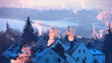 Abgase aus Schornsteinen auf schneebedeckten Dächern werden von der niedrigstehenden Sonne angestrahlt. | Bild: picture-alliance / Helga Lade Fotoagentur GmbH, Ger | N. Fischer