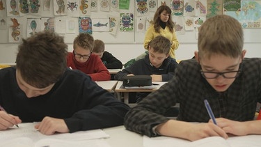 In einer Schulklasse sitzen Jugendliche beim Schreiben eines Texts am Schreibtisch. Im Hintergrund steht die Lehrerin der Brückenklasse an der Realschule Freiham.  | Bild: BR