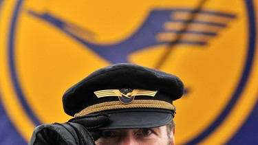 Lufthansa-Tarifstreit mit Pilotenvereinigung Cockpit dauert an; Symbolbild: Pilot steht vor Lufthansa-Logo | Bild: picture-alliance/dpa/Boris Roessler