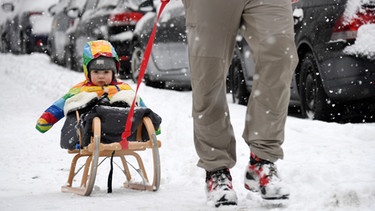 Ein Mann zieht seine Tochter Carla Maria in München (Bayern) auf einem Schlitten über einen verschneiten Bürgersteig.  | Bild: picture-alliance/dpa/Tobias Hase