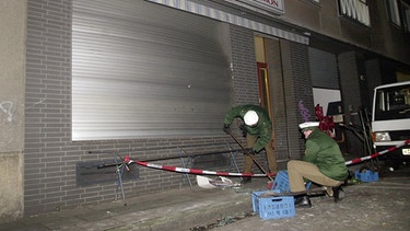 Polizeibeamte kehren nach einen Bombenanschlag auf ein Lebensmittelgeschäft am 19.01.2001 in der Probsteigasse in Köln herumliegende Scherben zusammen.  | Bild: picture-alliance/dpa