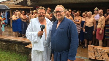 Zwei Männer in Bademänteln stehen vor einer Sauna. Im Hintergrund sind mehrere Menschen in Handtücher eingewickelt zu sehen. | Bild: BR-Studio Franken/Heiner Gremer