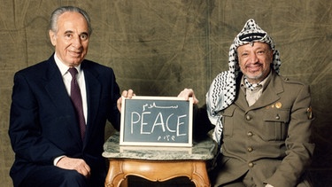 Palästinenserführer Jassir Arafat (r) und Israels Außenminister Shimon Peres halten am 12.12.1994 gemeinsam eine Tafel mit der Aufschrift "Peace" (dt.: Frieden) in den Händen. Arafat und Peres erhielten 1994 gemeinsam mit dem israelischen Ministerpräsidenten Jitzchak Rabin den Friedensnobelpreis | Bild: dpa/picture-alliance