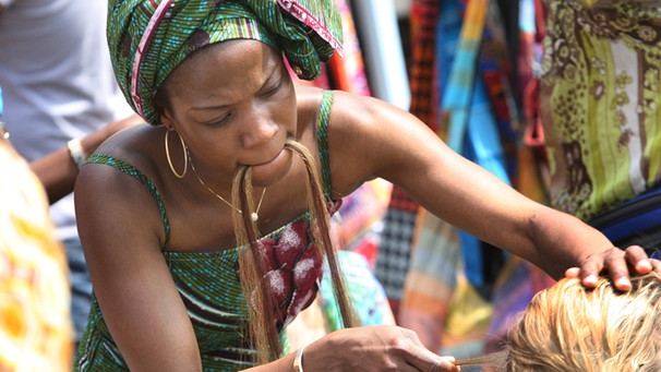 Eine Afrikanerin flechtet einer Besucherin Zöpfe ins Haar. | Bild: picture-alliance/dpa