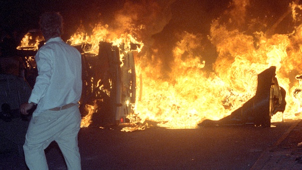 Ein Mann steht am 27.08.1992 vor einem brennenden Pkw auf einer Straße am zentralen Asylbewerberheim von Mecklenburg-Vorpommern in Rostock-Lichtenhagen | Bild: picture-alliance/dpa