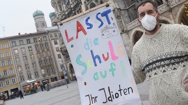 Ein Mann steht mit einem Schild mit der Aufschrift "Lasst die Schule auf - Ihr Idioten" vor dem Münchner Rathaus am Marienplatz. Archivbild vom 10.12.2020- | Bild: picture-alliance/dpa