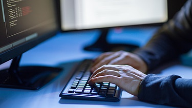 Hände tippen auf einer Tastatur vor einem Monitor mit Programmcode auf dem Bildschirm.  | Bild: picture alliance / Zoonar | lev dolgachov