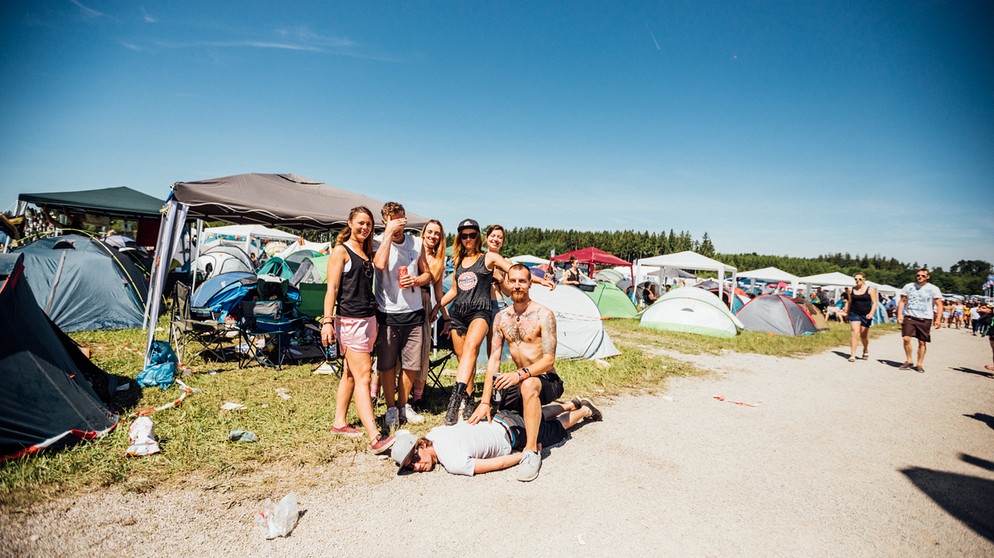 Eine glückliche Gruppe von Festival-Großwildjägern posiert mit ihrer Beute: ein Typ mit Sonnenbrille und weißem Shirt, der bäuchlings im Kiesweg am Campingplatz liegt. | Bild: BR/Johanna Schlüter