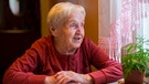 Eine alte Dame, etwa 90 Jahre alt, blickt mit einem Lächeln im Gesicht aus dem Fenster, sie sitzt davor an einem Tisch. | Bild: mauritius-images