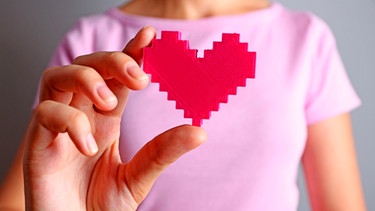 Frauenhand hält ein magentafarbenes Herz vor sich. Sie trägt ein rosafarbenes T-Shirt. | Bild: mauritius-images