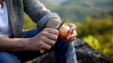 Hände eines Mannes schneiden unter freiem Himmel einen Apfel auf | Bild: mauritius images